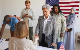 Multi-ethnic group of people at polling station decorated with American flags on election day, focus on smiling senior woman shaking hands with voting official, copy space